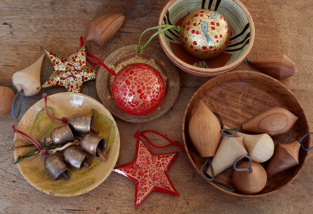 a tabletop filled with artisan christmas baubles in wooden and ceramic bowls including hand-painted red stars, wooden baubles and cow bells