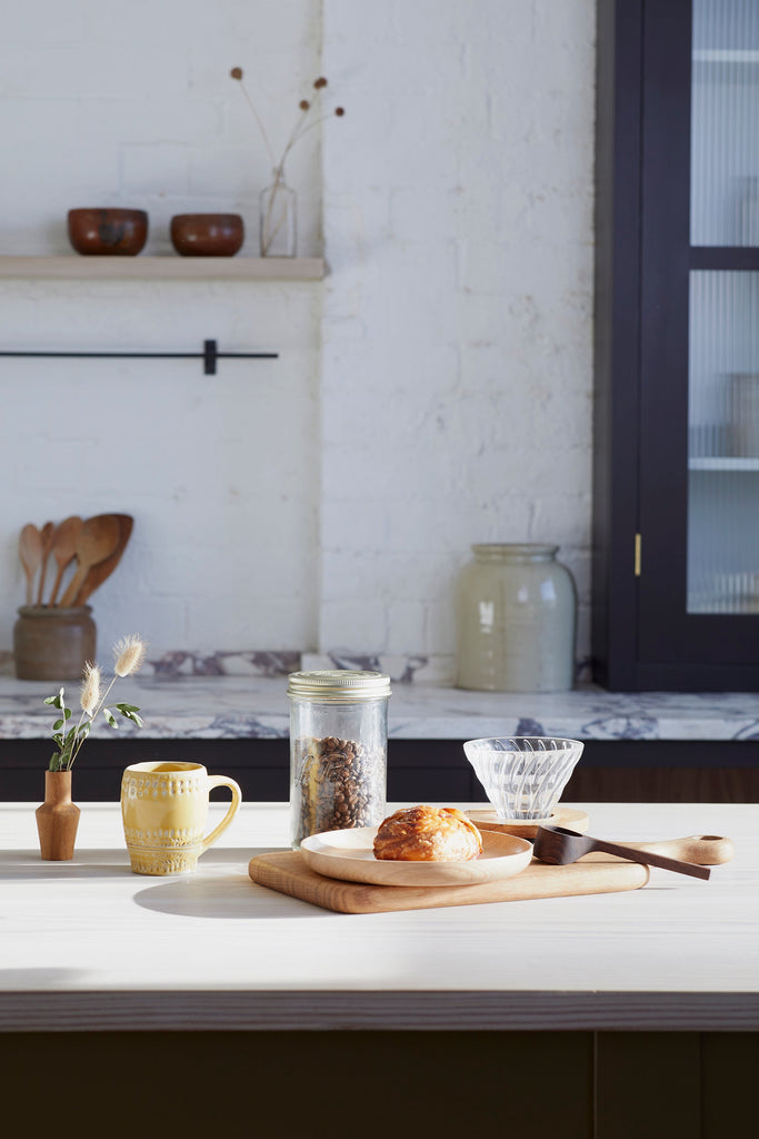 a coffee and breakfast scene with a mug, wooden coffee scoop, glass jar of coffee beans and v60 pourover